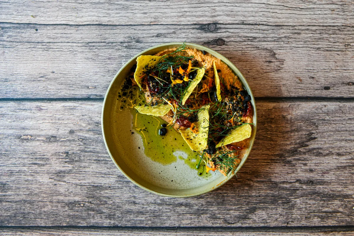 Hummus pomidorowy z ziemią z oliwek i chrupiącą tortillą, skropiony oliwą rozmarynową, podany w ceramicznym półmisku.