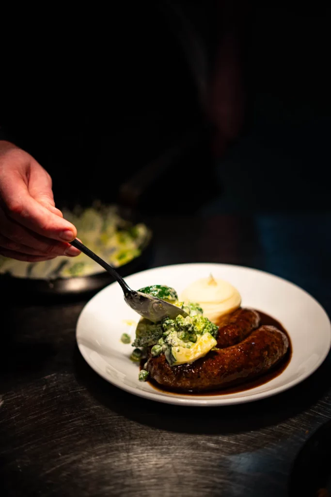 Biała kiełbasa z dziczyzny z sosem piwnym oraz gulaszem z zielonych warzyw i kremowymi ziemniakami podane na ceramicznym talerzu.