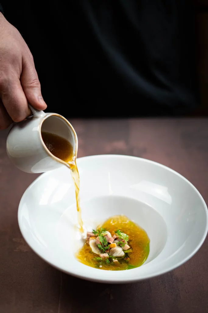 Rosół z pieczonej kaczki i uszkami z karmelizowanej szalotki oraz olejem lubczykowym podany w ceramicznym talerzu.